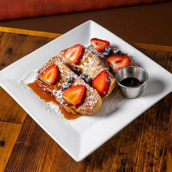 French toast with strawberries, blueberries, powdered sugar, and syrup on a white plate.