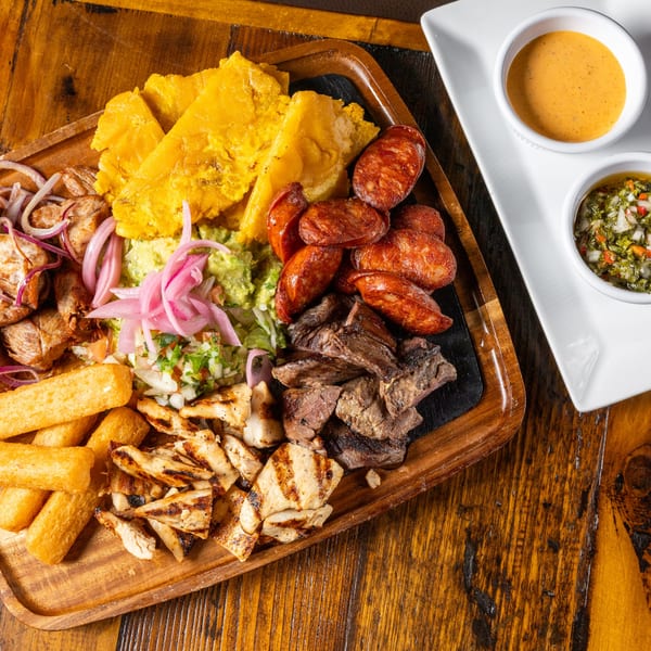 A wooden platter filled with grilled meats, plantains, guacamole, and pickled onions, with two dipping sauces.
