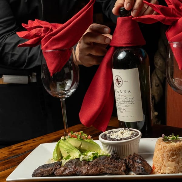 Waiter serving a steak dinner with avocado, rice, and beans, alongside a bottle of Malbec wine.