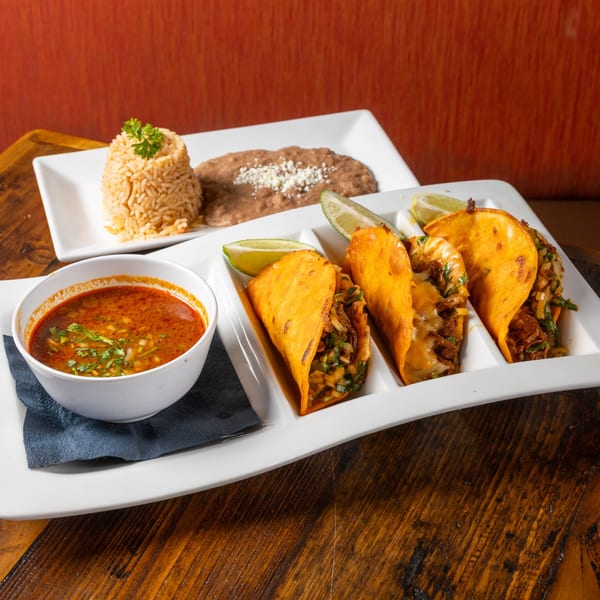 Three birria tacos with lime wedges, a bowl of consommé, and a plate of rice and beans.