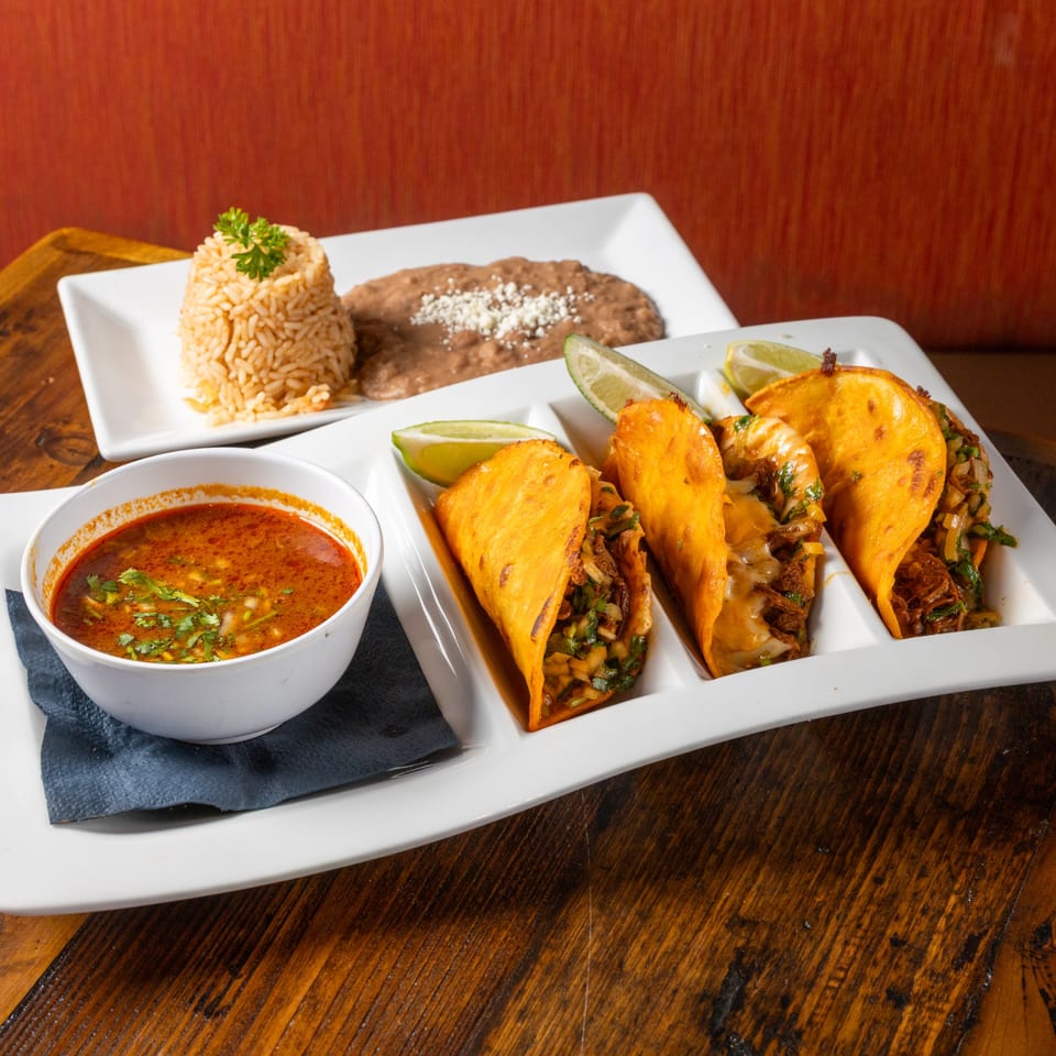 Three birria tacos with lime wedges, a bowl of consommé, and a plate of rice and beans.