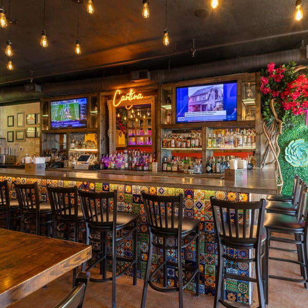 A vibrant bar with colorful tiled facade, liquor bottles, TVs, and a "Cantina" neon sign.
