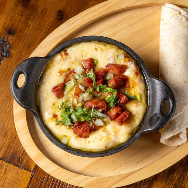 Melted cheese with chorizo, onions, and cilantro in a black skillet, with tortillas on a wooden tray.