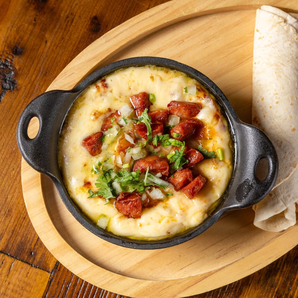 Melted cheese with chorizo, onions, and cilantro in a black skillet, with tortillas on a wooden tray.