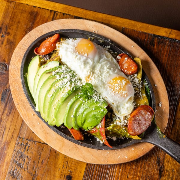 A sizzling skillet with fried eggs, sliced avocado, chorizo, and crumbled cheese on a wooden board.