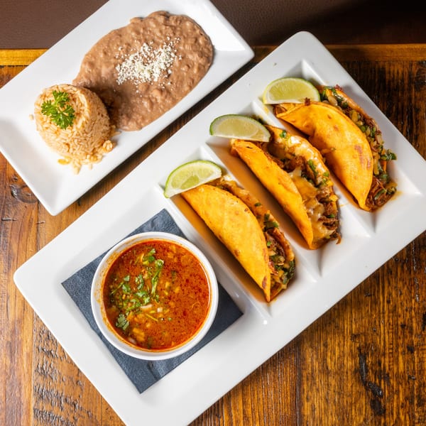 Frida's Mexican Grill birria tacos with consommé, rice, and refried beans on a wooden table.