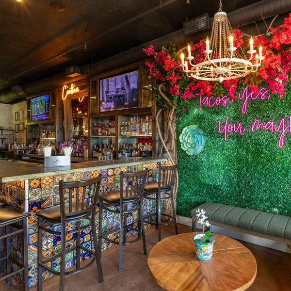 Vibrant bar interior with colorful tiled counter, neon sign, and lush floral decor.