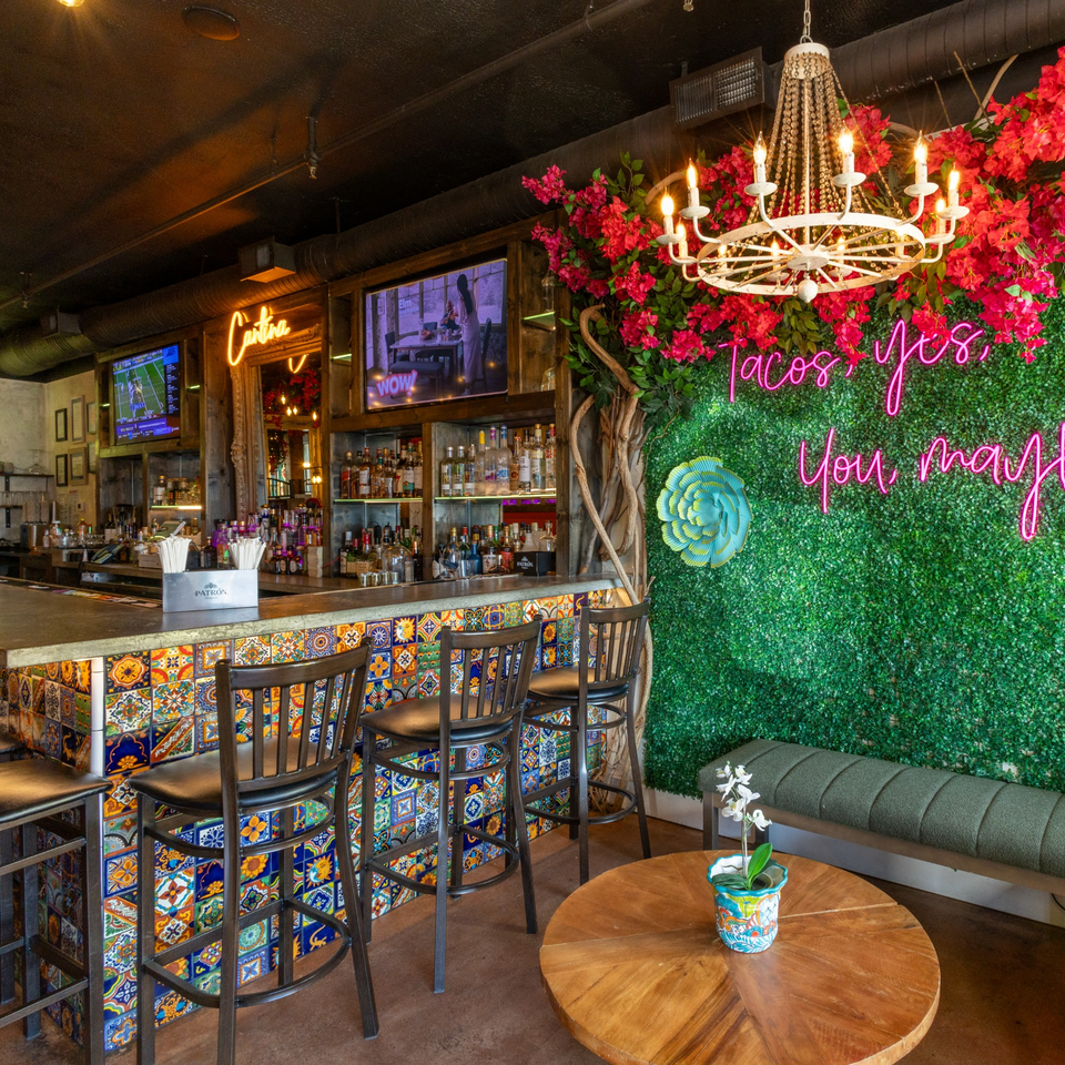 Vibrant bar interior with colorful tiled counter, neon sign, and lush floral decor.