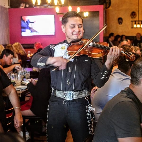 A mariachi musician in a black suit and silver belt plays violin for diners at Frida's Mexican Grill.