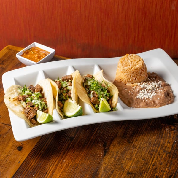 Three Frida's Mexican Grill carne asada tacos with lime, rice, and refried beans on a white plate.