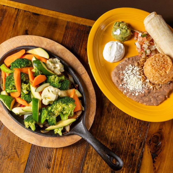 Sizzling fajita vegetables in a cast iron pan next to a Mexican plate with rice, beans, and tortillas.