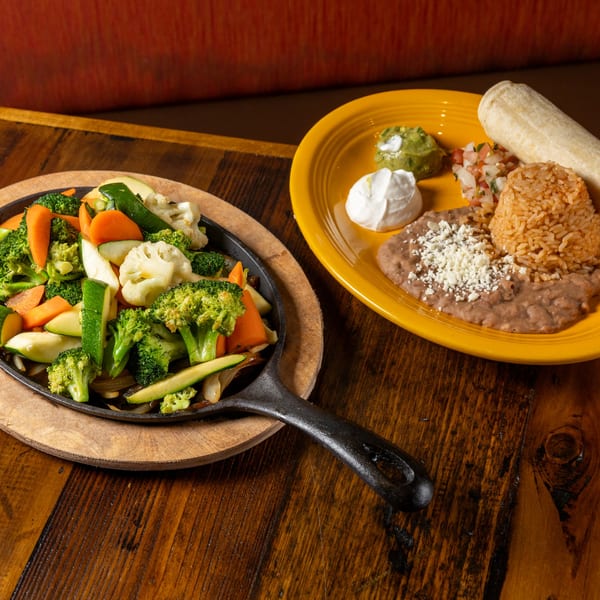 Sizzling fajita vegetables in a cast iron pan next to a plate of Mexican rice, beans, and tortilla.