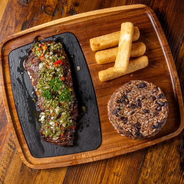 Grilled steak with chimichurri, yucca fries, and rice with black beans on a wooden tray.