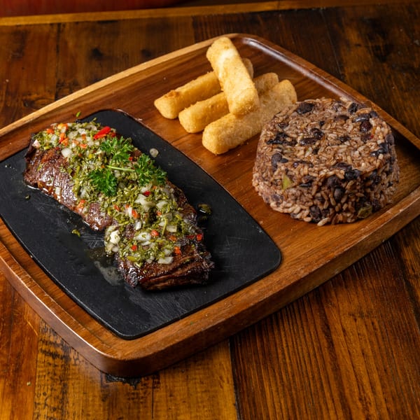 Grilled steak with chimichurri, black bean rice, and fried yuca sticks on a wooden tray.