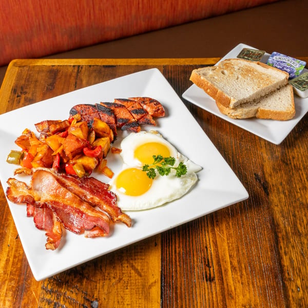 Breakfast plate with fried eggs, bacon, sausage, potatoes, and toast with jelly on a wooden table.