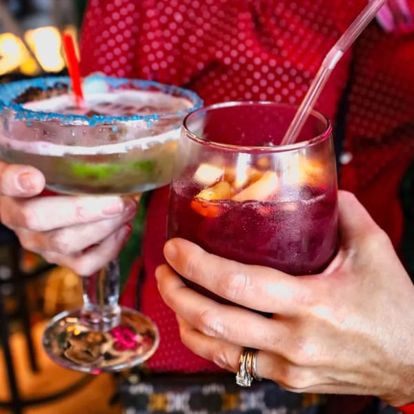 Two people clinking glasses, one with a blue-rimmed margarita, the other with red sangria.