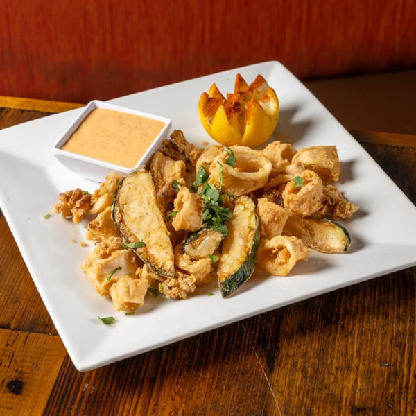 Crispy fried calamari and zucchini with a side of creamy dipping sauce and a lemon garnish.