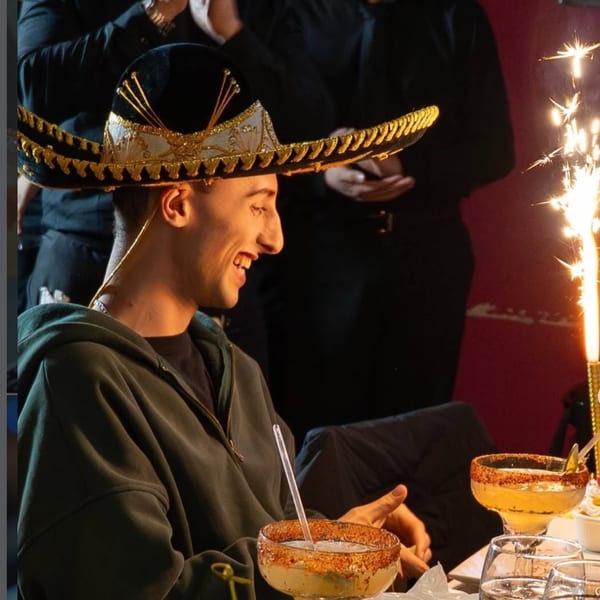 A smiling man wearing a sombrero, with two margaritas and a sparkler on the table.