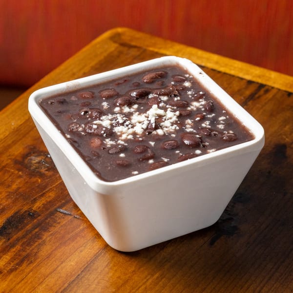 A white square bowl of Frida's Mexican Grill's black beans topped with crumbled white cheese.