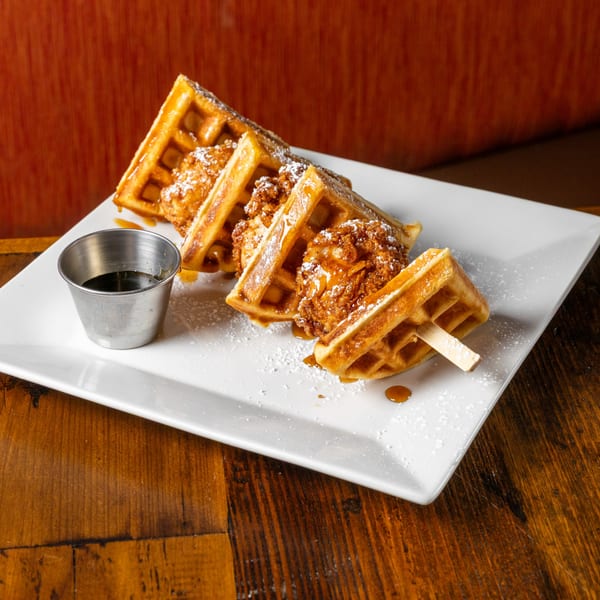 Chicken and waffles on skewers, drizzled with syrup and powdered sugar, served with a side of syrup.