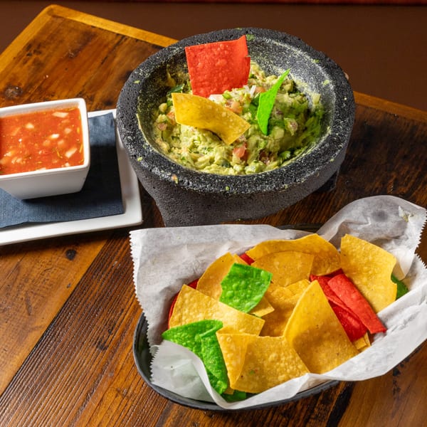 Guacamole in a molcajete, salsa, and a basket of colorful tortilla chips on a wooden table.