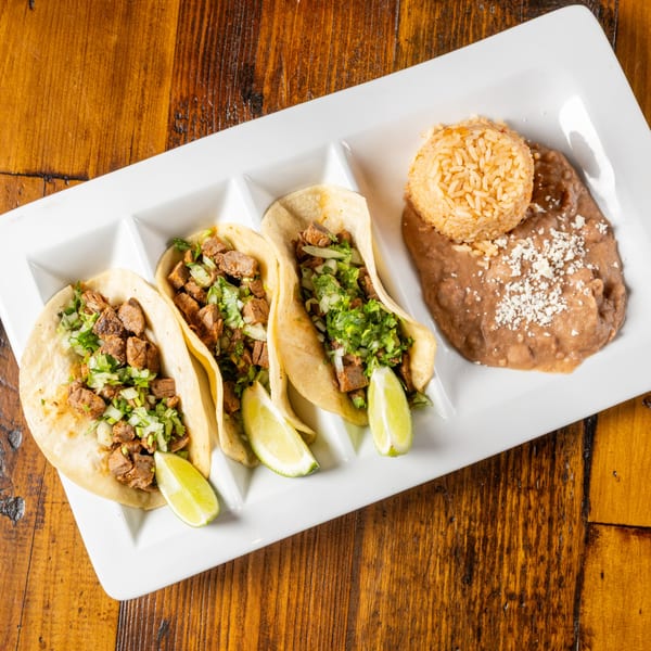 Three street tacos with meat, onions, cilantro, and lime wedges, served with rice and refried beans.